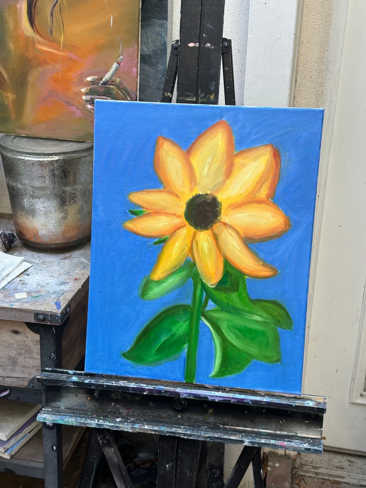 Oil painting of a yellow sunflower against a bright blue background, resting on a studio easel.